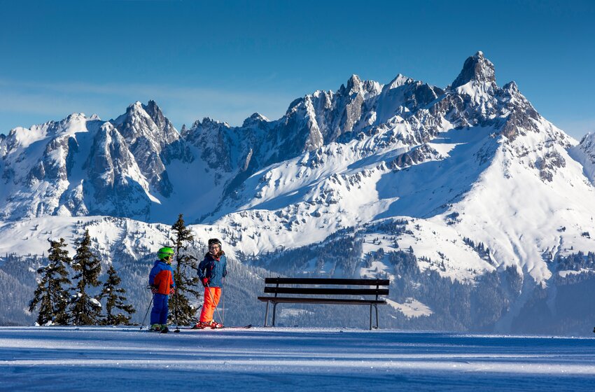 Two kids on skis in front of a bench with stunning snowy alpine mountain backdrop. | © Salzburger Sportwelt Amadé GmbH