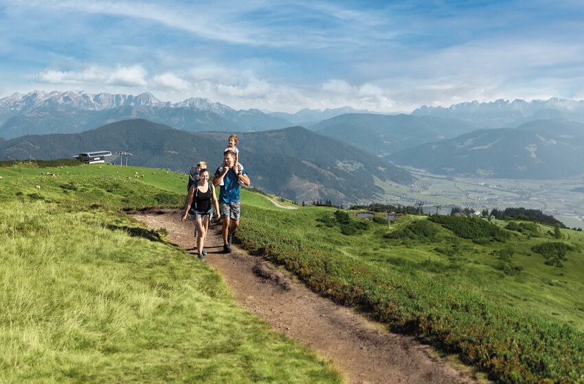 Family with two children hikes along a mountain trail through green alpine meadows with a wide Alpine panorama. | © Snow Space Salzburg