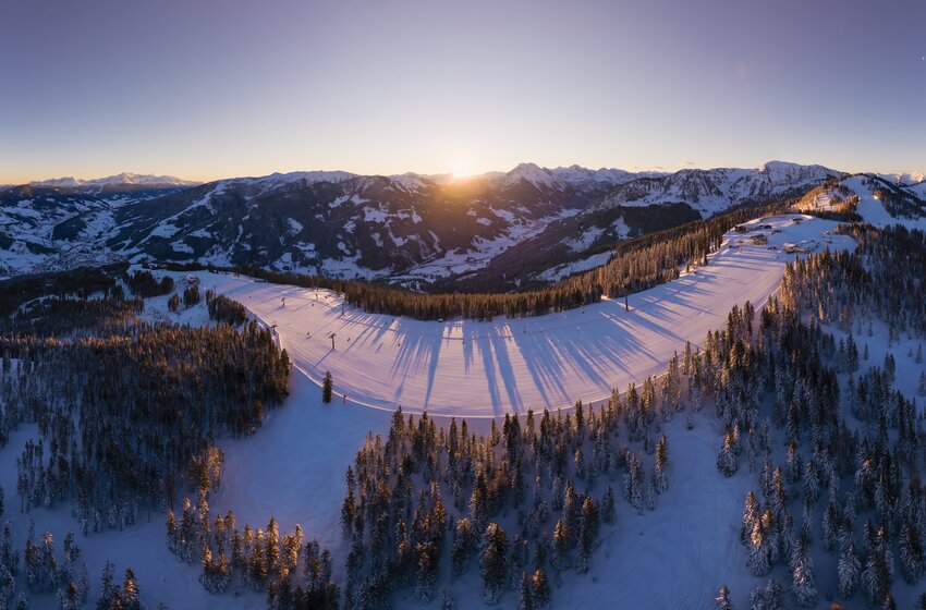 Luftaufnahme verschneiter Skipisten und Wälder im Morgenlicht mit tiefen Schatten vor einem weiten Alpenpanorama. | © Snow Space Salzburg 