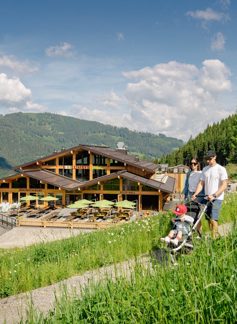 Family with child in stroller walks across alpine meadow to modern wooden mountain hut with sun terrace. | © SHUTTLEBERG GmbH & Co KG
