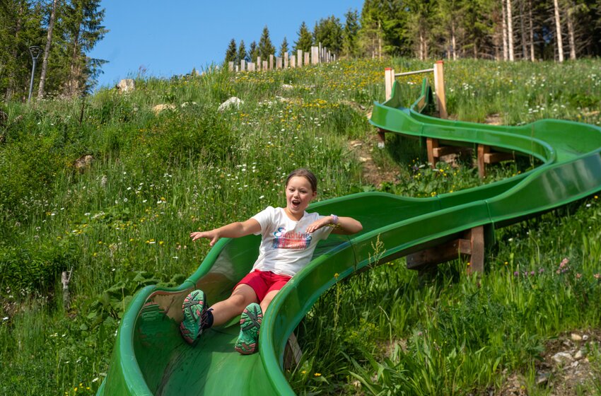 Laughing girl slides on green curved slide through flower-covered slope