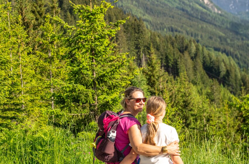 Mother and daughter sit on wooden bench facing forested sunlit mountain | © SHUTTLEBERG GmbH & Co KG
