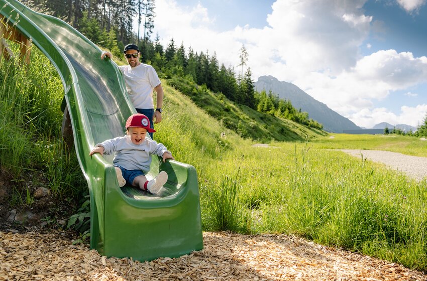 Toddler laughing while sliding on green slide, father stands behind | © SHUTTLEBERG GmbH & Co KG