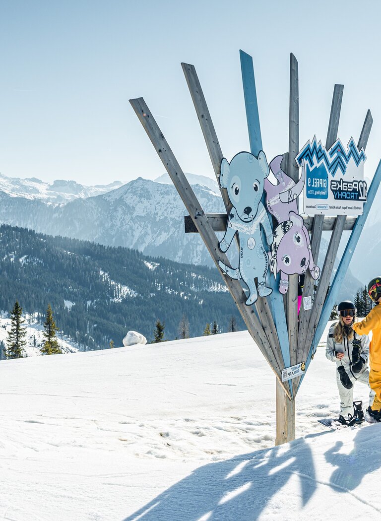 Kids in colorful winter outfits with snowboards at Familypark mascot sign, mountain scenery in background. | © SHUTTLEBERG GmbH & Co KG