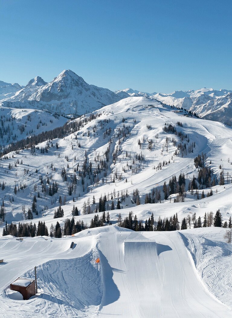 View across a groomed snow park with kickers and rails in front of snow-covered mountains beneath a clear blue sky. | © SHUTTLEBERG GmbH & Co KG