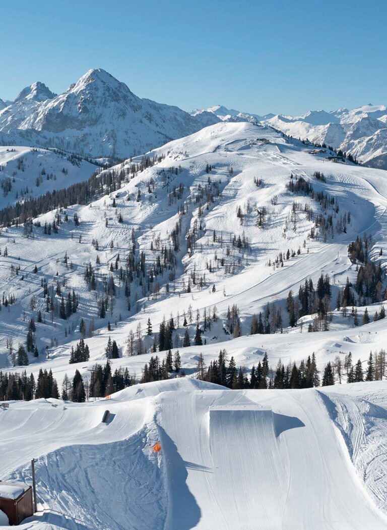 Blick über einen präparierten Snowpark mit Kickern und Rails vor verschneiten Bergen unter klarem blauem Himmel. | © SHUTTLEBERG GmbH & Co KG