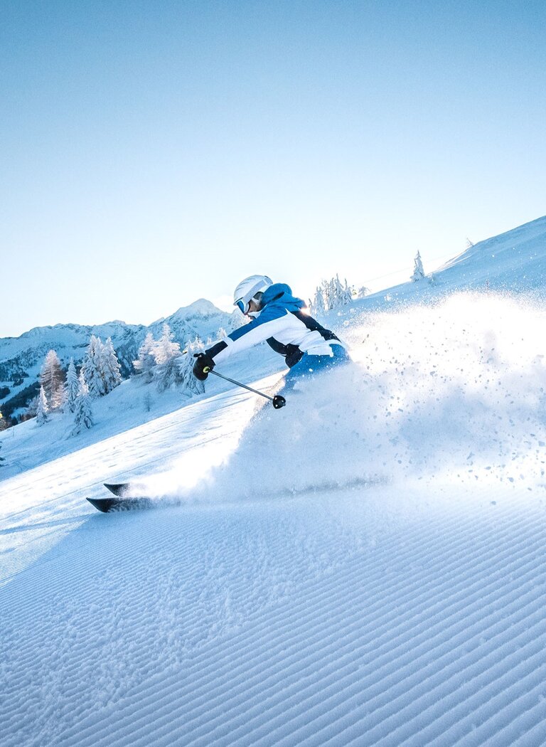 Skifahrer wirbelt Pulverschnee auf sonniger Piste in Ski amadé auf | © Ski amadé GmbH Ein Skifahrer zieht bei Sonnenlicht Schwünge durch frischen Schnee und wirbelt Pulverschnee auf einer Piste in Ski amadé auf. | © Ski amadé GmbH