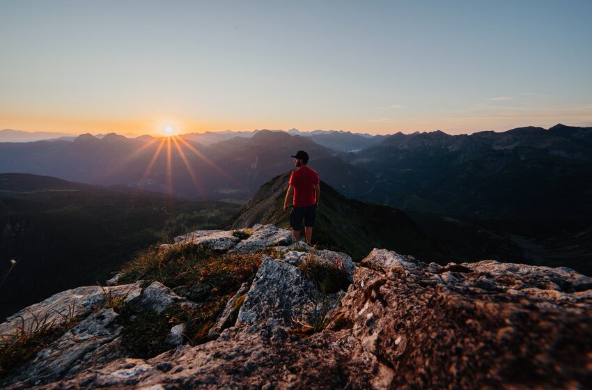 A hiker stands on a rocky summit watching the sunrise over a vast Alpine mountain panorama. | © Zauchensee Liftgesellschaft Benedikt Scheffer GmbH