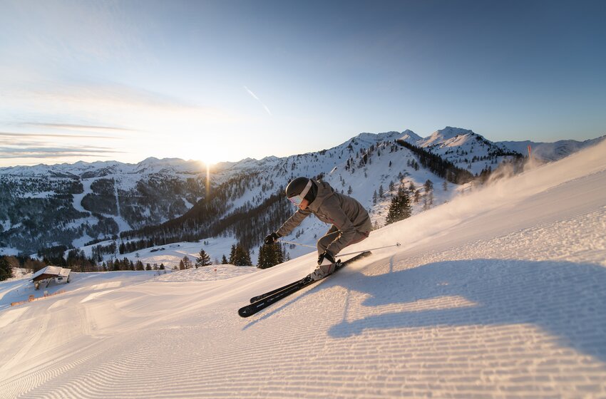 Ein Skifahrer zieht einen Schwung auf einer präparierten Piste vor verschneiten Bergen bei tief stehender Sonne. | © Christian Schartner